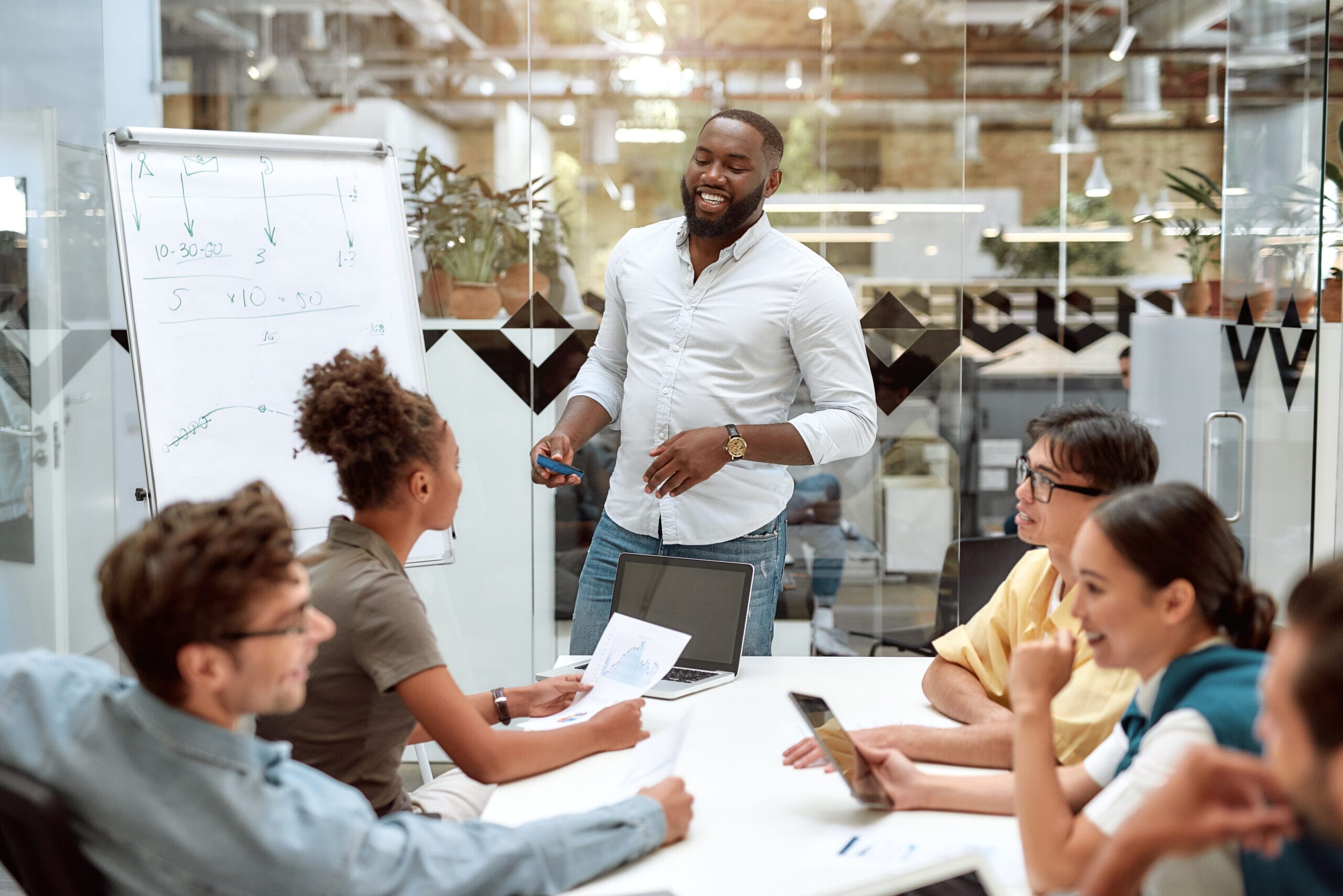 Young African American businessman pointing at white blackboard and smiling while working with team in the creative office